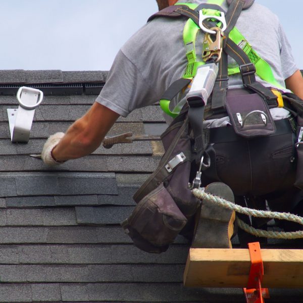 A professional roofer, equipped with a safety harness and tool belt, installs asphalt shingles on a steep residential roof under a cloudy Weston, TX sky. The close-up image shows hand tools in use, secure anchor points, and clean shingle alignment, emphasizing workmanship and safety practices. SCR, Inc.—a trusted Weston roofing company—offers high-quality residential and commercial roof installations and repairs. With over 25 years of experience, they’re known for attention to detail, insurance expertise, and 24/7 emergency response. Call 972-839-6834 for roofing help in Weston, TX.